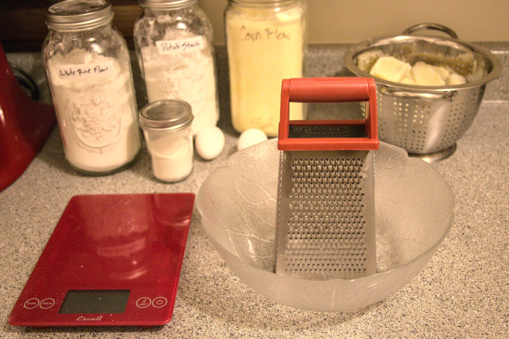 A countertop with a scale, a large bowl with a grater sitting inside, a colander of cooked potatoes, and jars of flour.