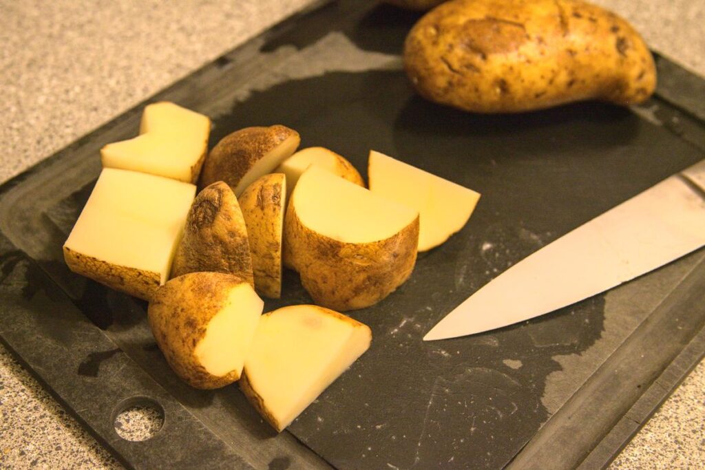 Potatoes cut into large chunks on a cutting board, accompanied by a knife and a whole potato