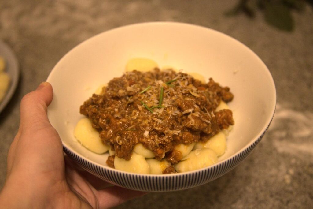 A hand holding a white bowl filled with gnocchi in a bolognese sauce.