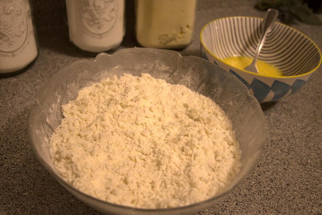 A large bowl filled with grated potato mixed with flour. A small bowl with two beaten eggs in it sits in the background.