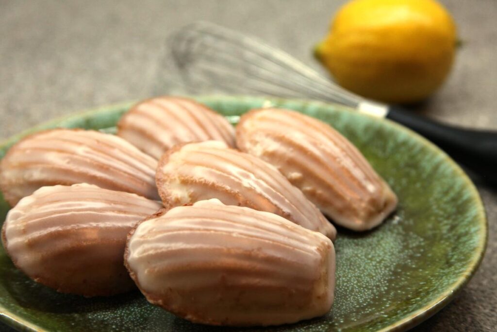 A green plate with six iced madeleines on it. In the background is a whisk and a lemon.