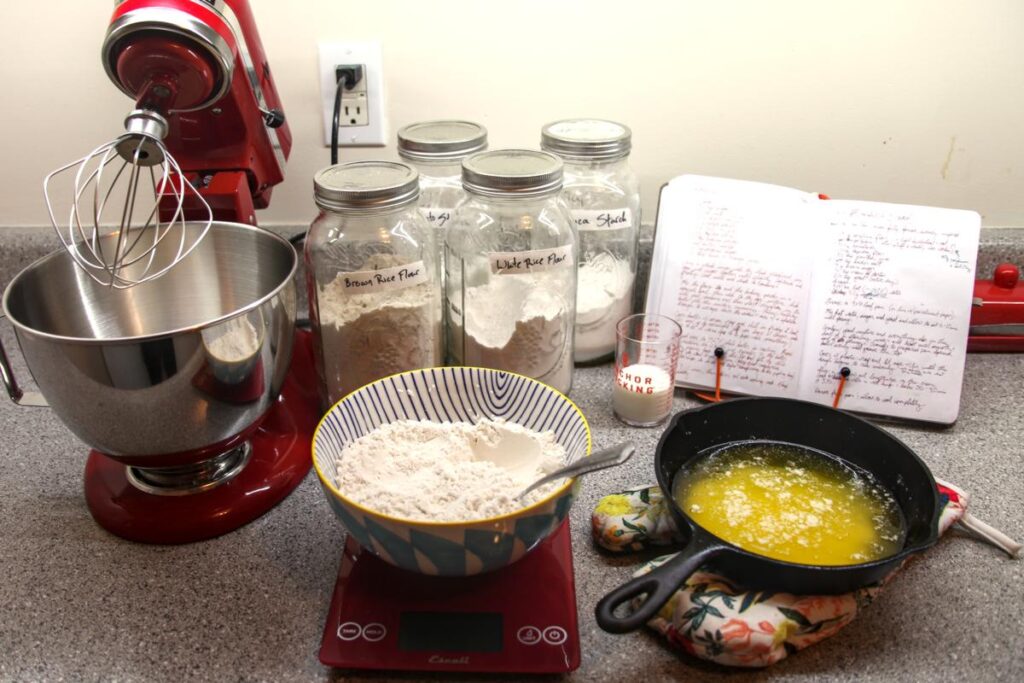 Prepared madeleine ingredients laid out on a countertop. There is a stand mixer, a bowl with the flour mix, and a small cast iron skillet with the melted butter. Behind them are jars of flour and a recipe book.
