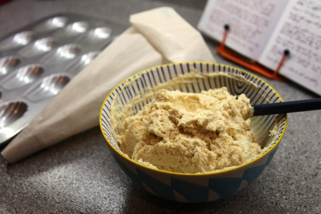 The chilled madeleine batter is in a bowl in the foreground. The batter is chunky. In the background are a piping bag, a madeleine tin, and a cookbook.