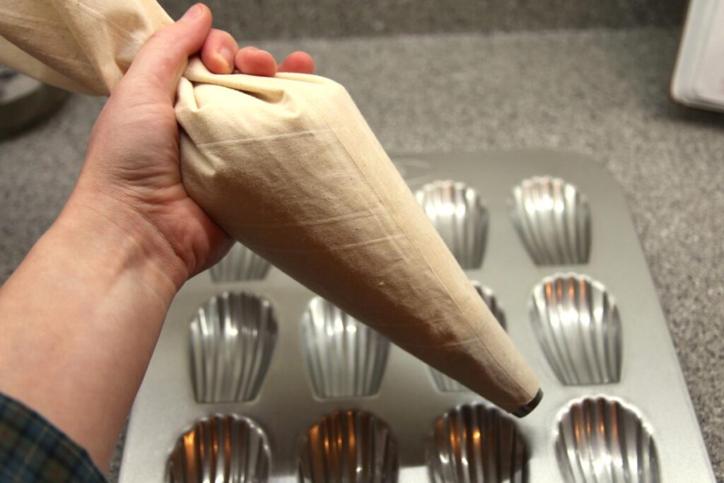 A hand holds a filled piping bag above an empty madeleine tin.