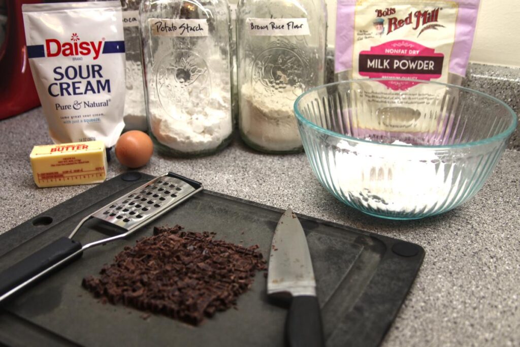 Flours, butter, an egg, and a cutting board with chopped chocolate and a grater on it are arranged on a countertop.