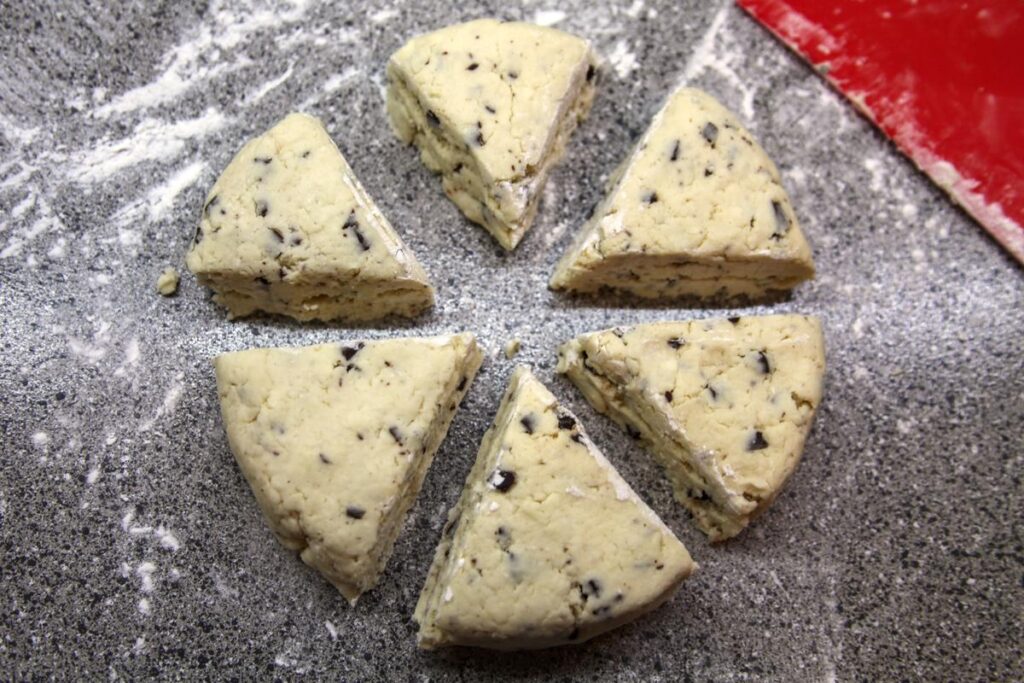 Six uncooked scones sit in a circle on a floured countertop. They have just been cut into sections from a round.