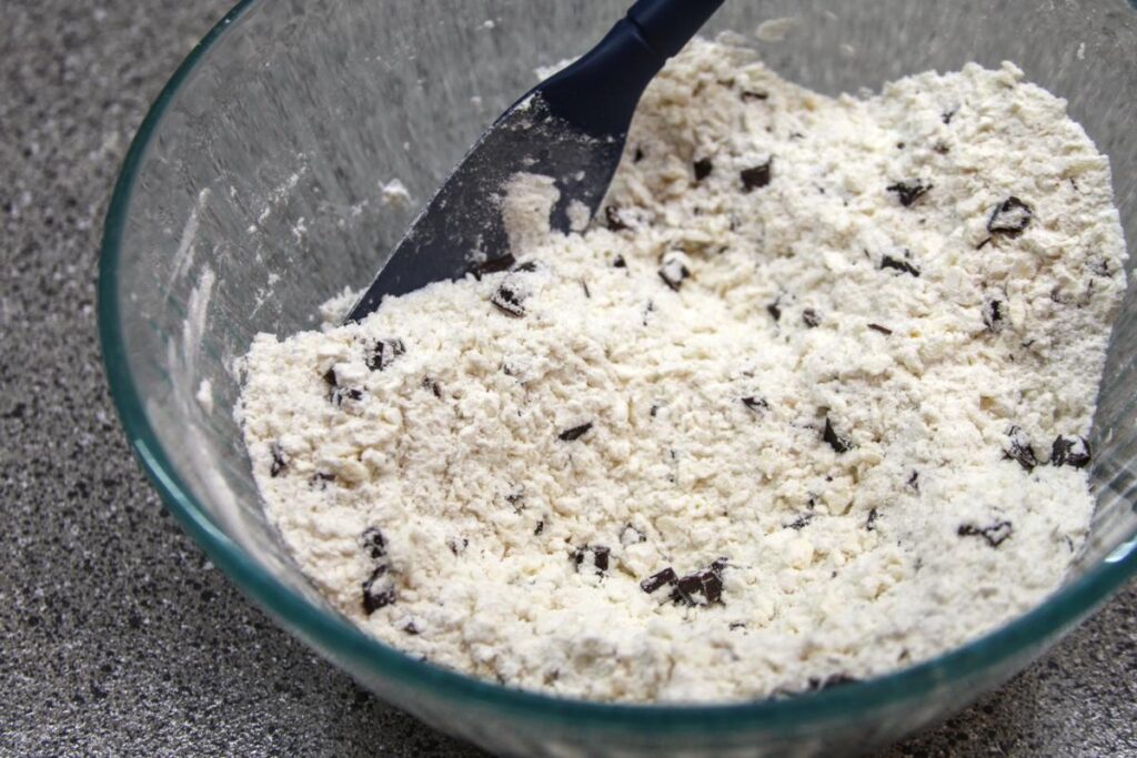 A bowl sits on a countertop with a spatula in it. The bowl contains flour mixed with grated butter and chocolate chunks.