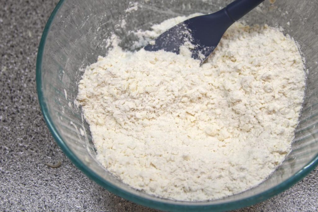 A bowl sits on a countertop with a spatula in it. The bowl contains flour mixed with grated butter.