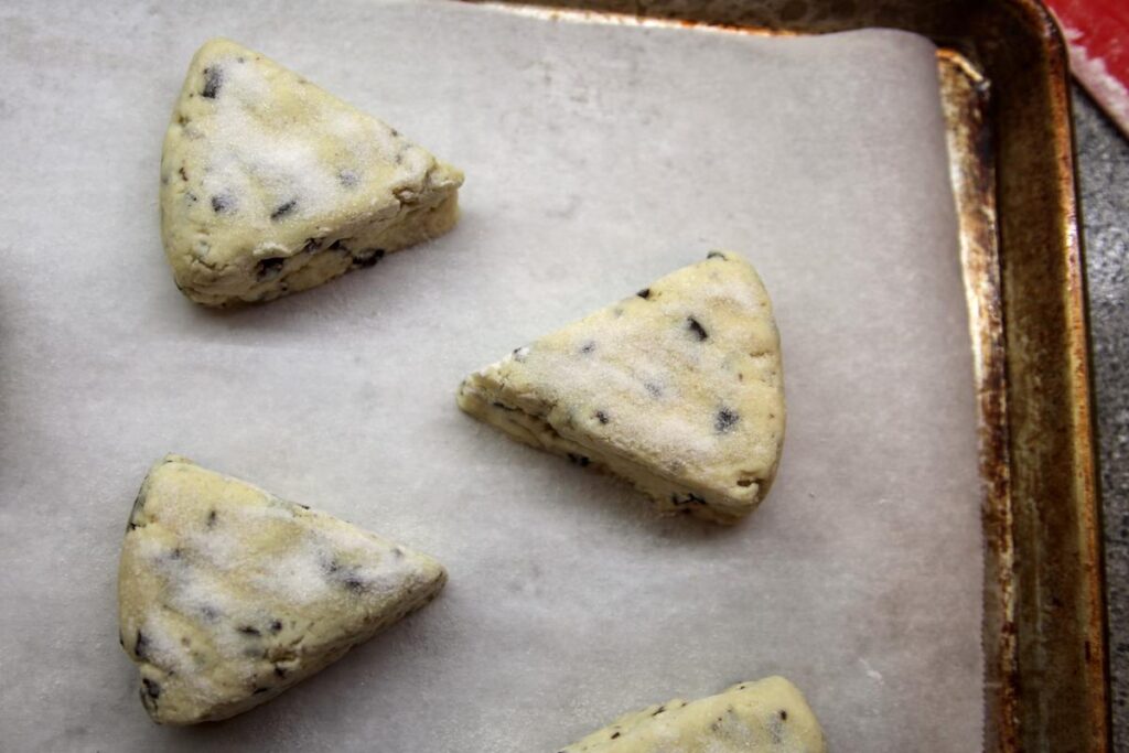 Several unbaked scones sit on a parchment paper lined baking sheet. The tops have been dusted with granulated sugar.