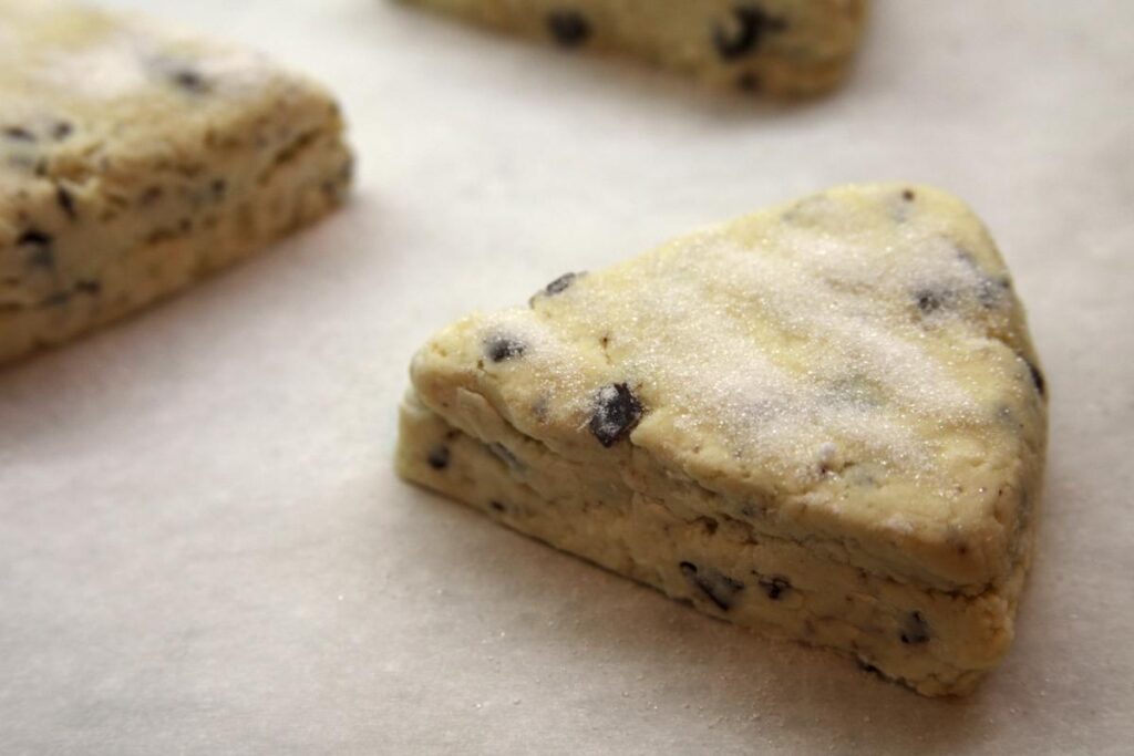 A close-up of an unbaked scone sitting on a parchment paper lined baking sheet. The top has been dusted with granulated sugar.