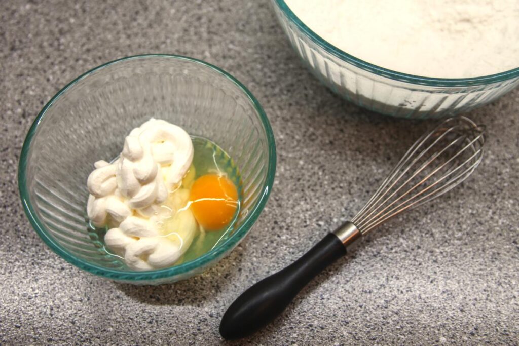 A bowl with sour cream and an egg cracked into it sits on the countertop next to a whisk and a bowl of flour.