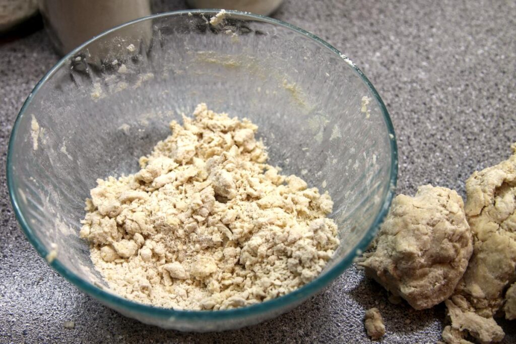 A clump of pie dough sits next to a clear glass bowl with dry flour and bits of pie dough at the bottom.