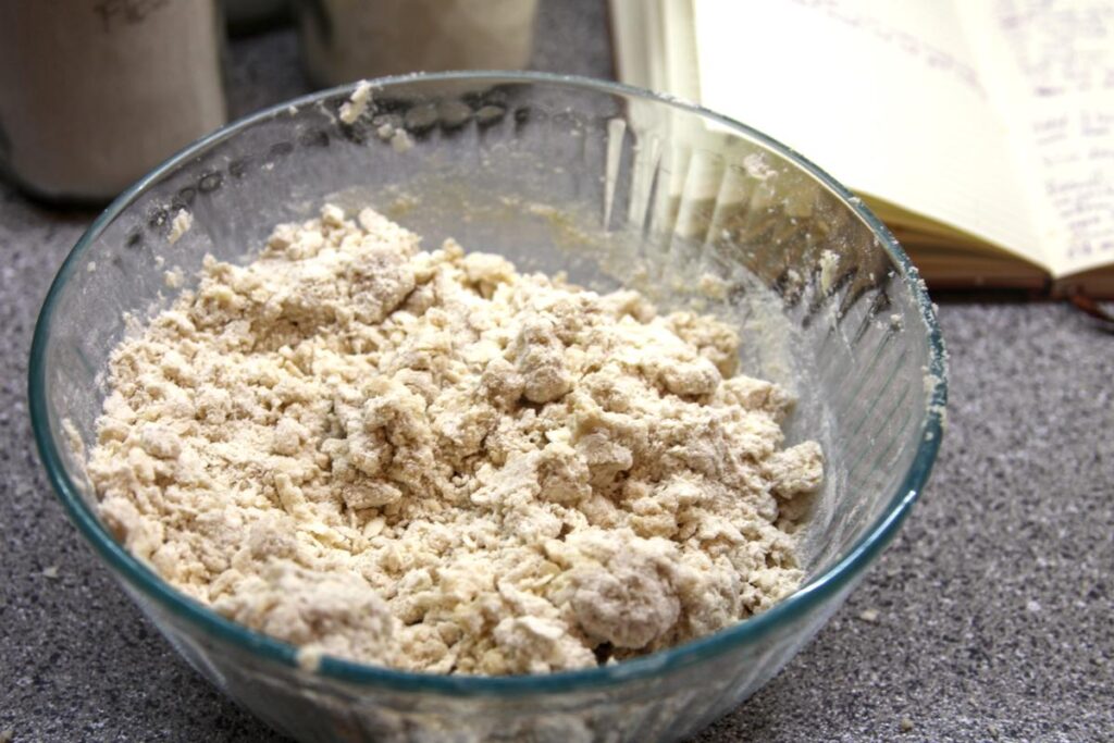 A clear glass bowl sits on a countertop. It is filled with a shaggy dough that looks not fully hydrated.