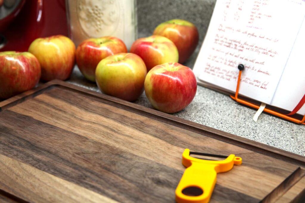 A wooden cutting board sits in the foreground with a vegetable peeler on it. In the background are seven apples and a recipe book on a stand.