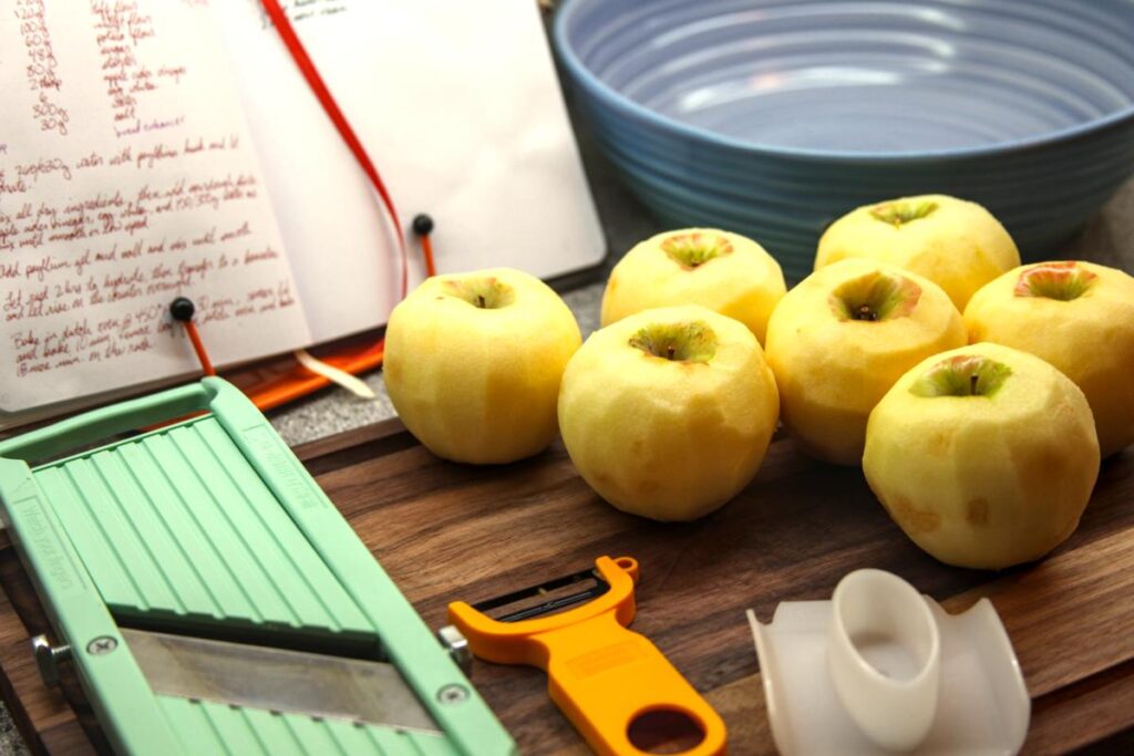 Peeled apples, a mandolin, and a vegetable peeler sit on a wooden cutting board. In the background is a purple bowl and a recipe book on a stand.