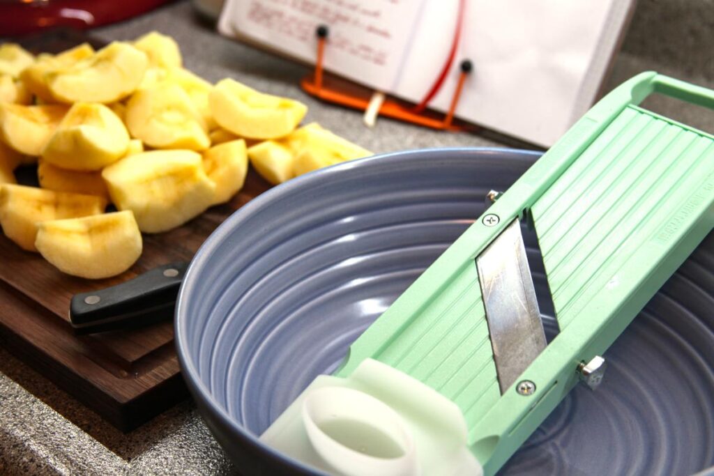 In the foreground is a purple bowl with a mandolin sitting in it. In the background is a wood cutting board with many peeled and cored apple quarters on it.