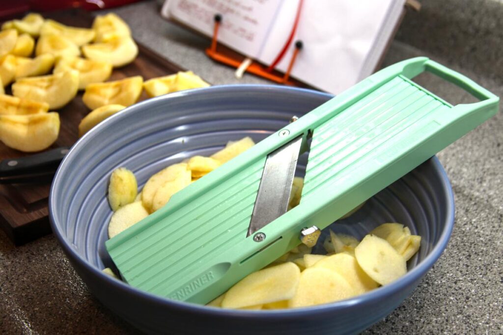 A purple bowl with a mandolin and sliced apples in it sits in the foreground. In the background is a wooden cutting board with many apple quarters on it.