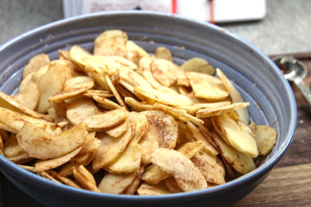 A purple bowl of apple slices. The slices are covered in a mix of flour, cinnamon, sugar, lemon juice, and sour cream.