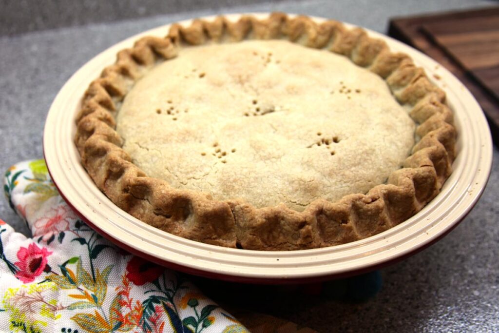 A large apple pie sits on a countertop. The pie plate is red with a white upper rim. Floral patterned hot pads sit next to it.