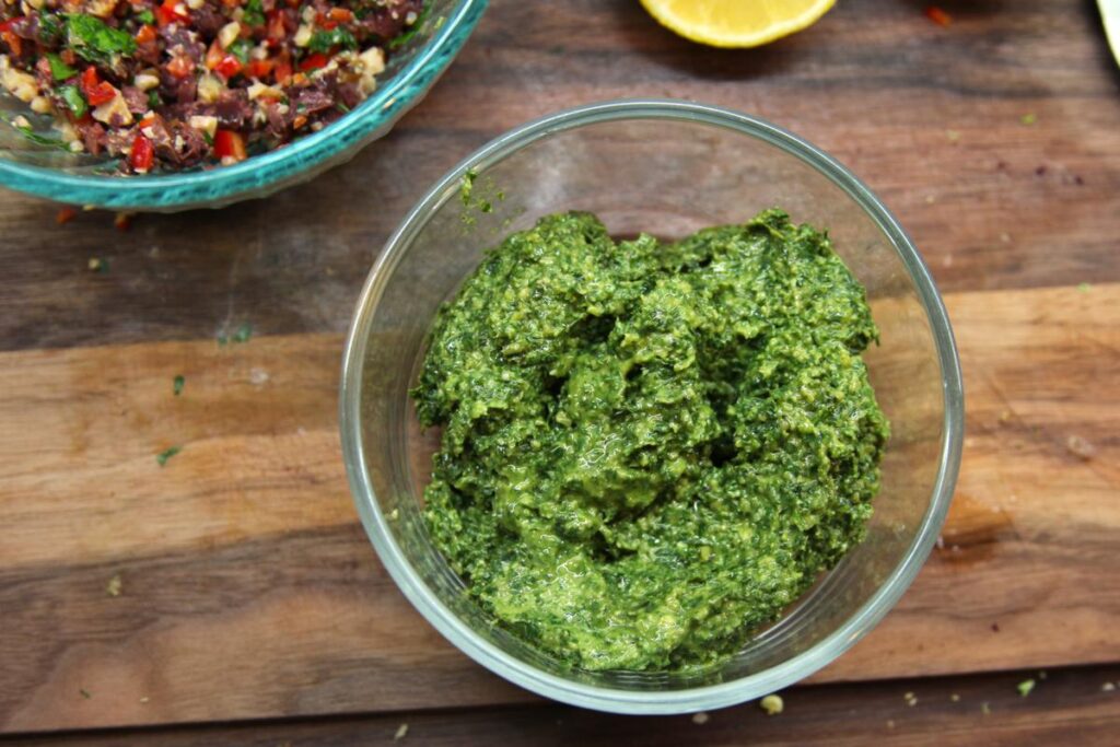 A glass bowl of basil pesto sits on a cutting board. In the background are a bowl of tapenade and a lemon.