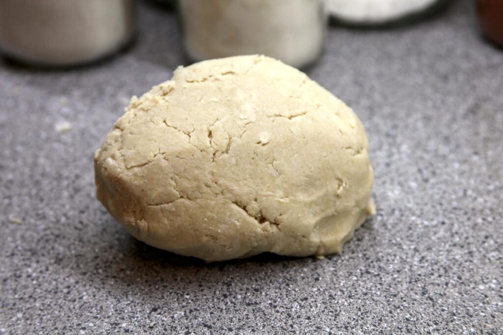 A rough ball of pie dough sits on the countertop in front of several glass jars. The dough surface is slightly cracked and butter can be seen swirled through the dough.