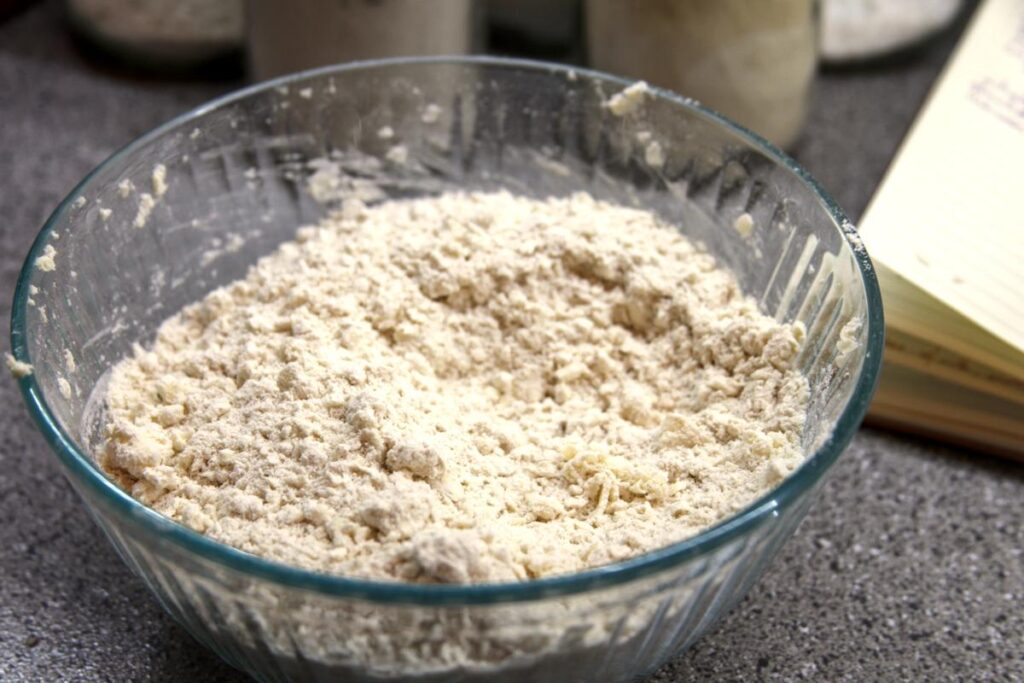A clear bowl filled with mixed together flour and grated butter sits on a countertop. The mixture is lumpy due to the intact butter pieces. There are jars and an open cookbook in the background.