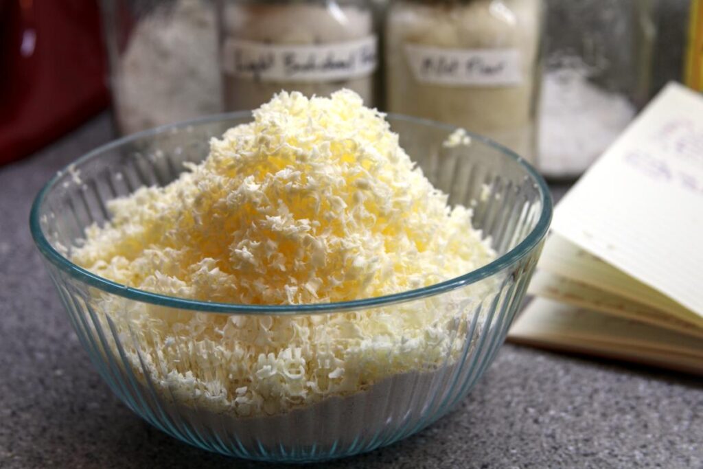 A clear bowl is filled with flour topped by a large mound of grated frozen butter. In the background are jars of flours and an open cookbook.