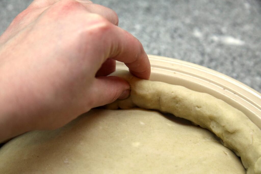A hand pinches the edge of a pie crust in between the thumb and middle and pointer fingers.