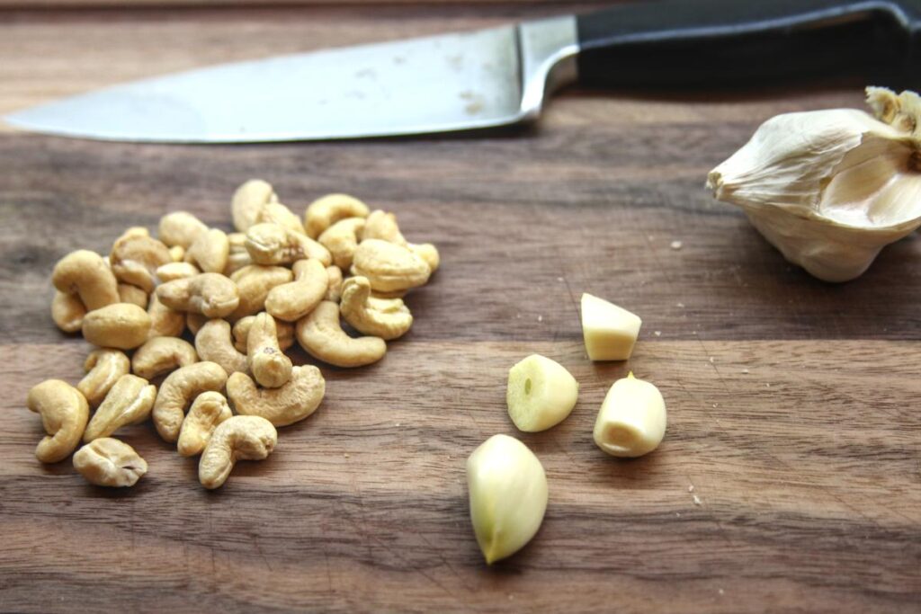 a handful of cashews, three garlic cloves, and a partially used head of garlic sit on a cutting board with a knife.