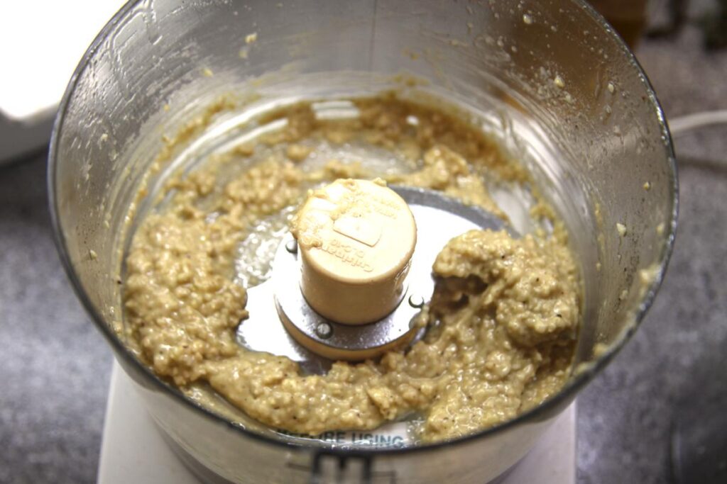A close-up of the bowl of a food processor containing a slightly chunky white-ish paste.