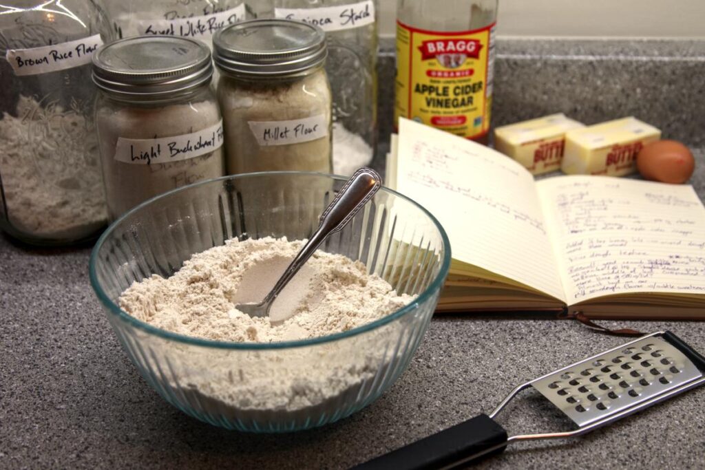 A bowl of flours with a spoon in it sits in the foreground, along with a handheld grater. In the background are jars of flower, a cookbook, a bottle of apple cider vinegar, two sticks of butter, and an egg.