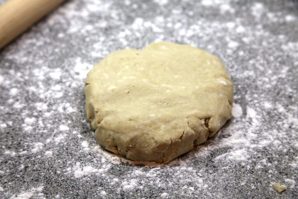 A flat disk of dough sits on a lightly floured countertop. Beside it is a french style rolling pin.