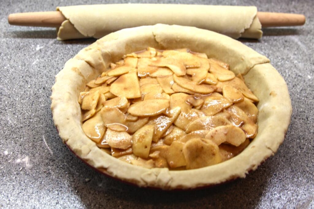 A pie plate filled with apple filling sits in the foreground. Behind it is a rolling pin with a sheet of pie crust wrapped around it, ready to cover the apples.