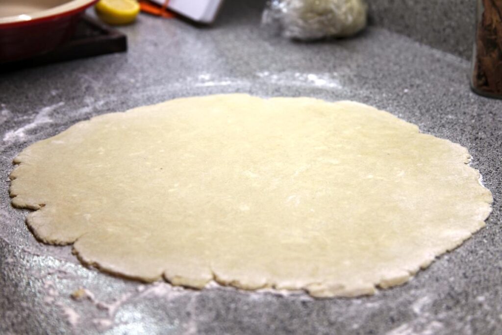 A large circle of rolled out pie dough rests on a lightly floured countertop. In the background are several indistinct objects.