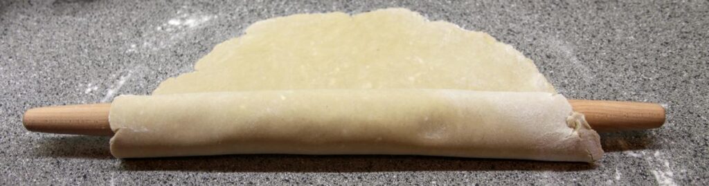 A long, thin picture shows a circle of pie dough partially rolled up around a long, French-style rolling pin.